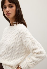 Cremfarbener Strickpullover mit Rippkragen und langen Ärmeln. Besitzt ein strukturiertes Muster und ist über einem kragenlosen Hemd mit Muster drapiert.
