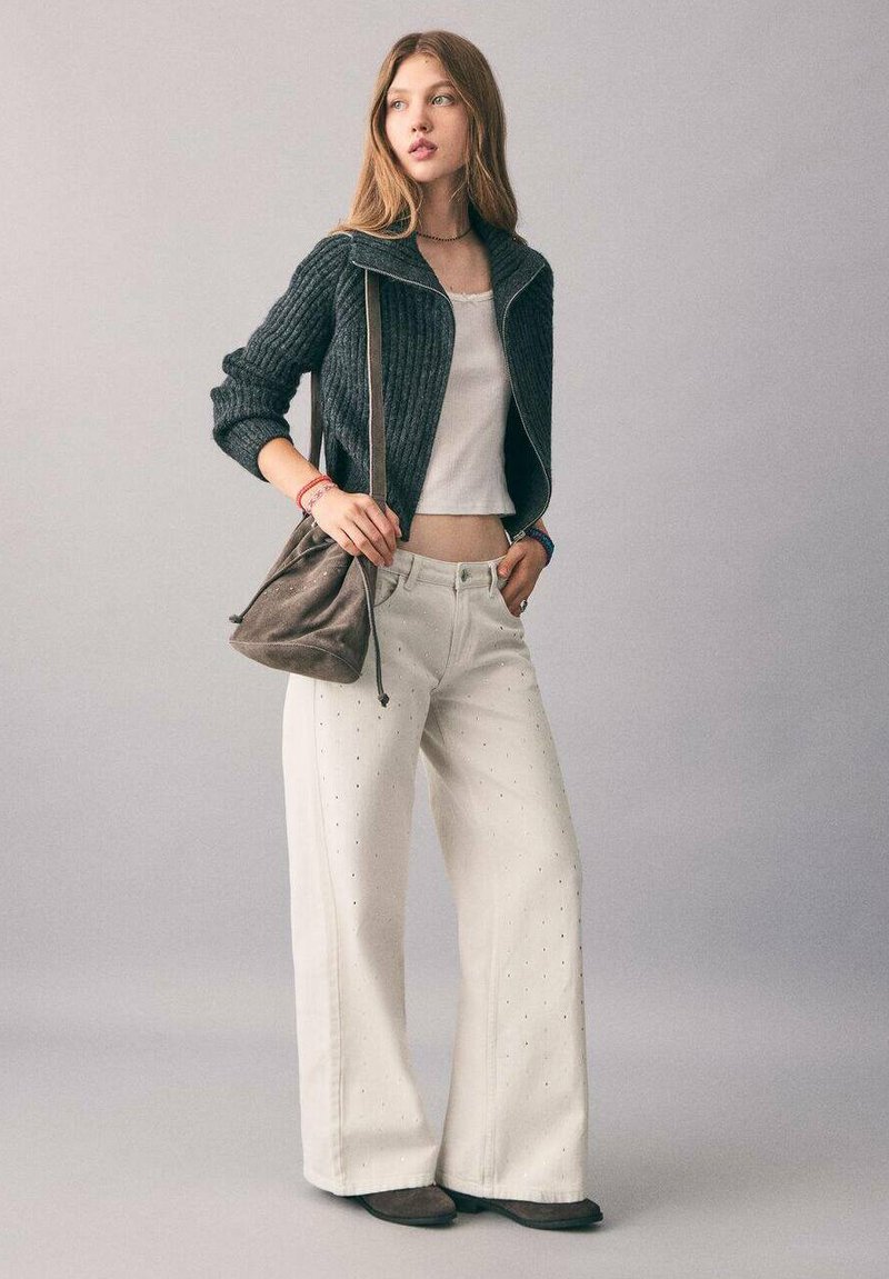 Chaqueta gris de cremallera con canalé, top corto blanco, pantalones anchos blancos con perforaciones, bolso cruzado marrón y zapatos planos marrones. Paleta de colores neutros.