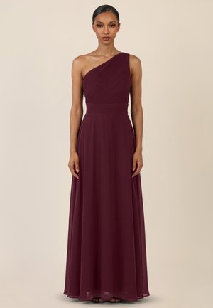 Frau steht in einem bodenlangen, einseitig schulterfreien bordeauxroten Kleid mit einem eng anliegenden, strukturierten Oberteil und einem fließenden Rock.