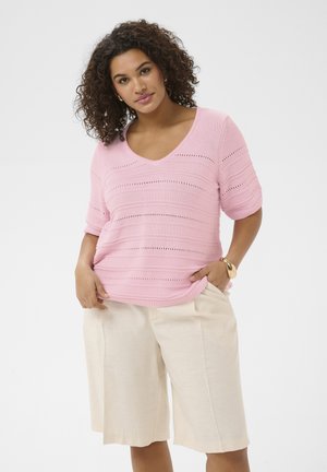 Femeie cu păr creț, purtând un tricou roz cu mâneci scurte din tricot și pantaloni scurți bej până la genunchi, cu o mână în buzunar și o brățară aurie.