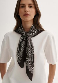 Czarny i biały jedwabny szalik w wzór paisley związany na szyi, kontrastujący z gładką, białą koszulą z krótkim rękawem. Miękka tekstura.