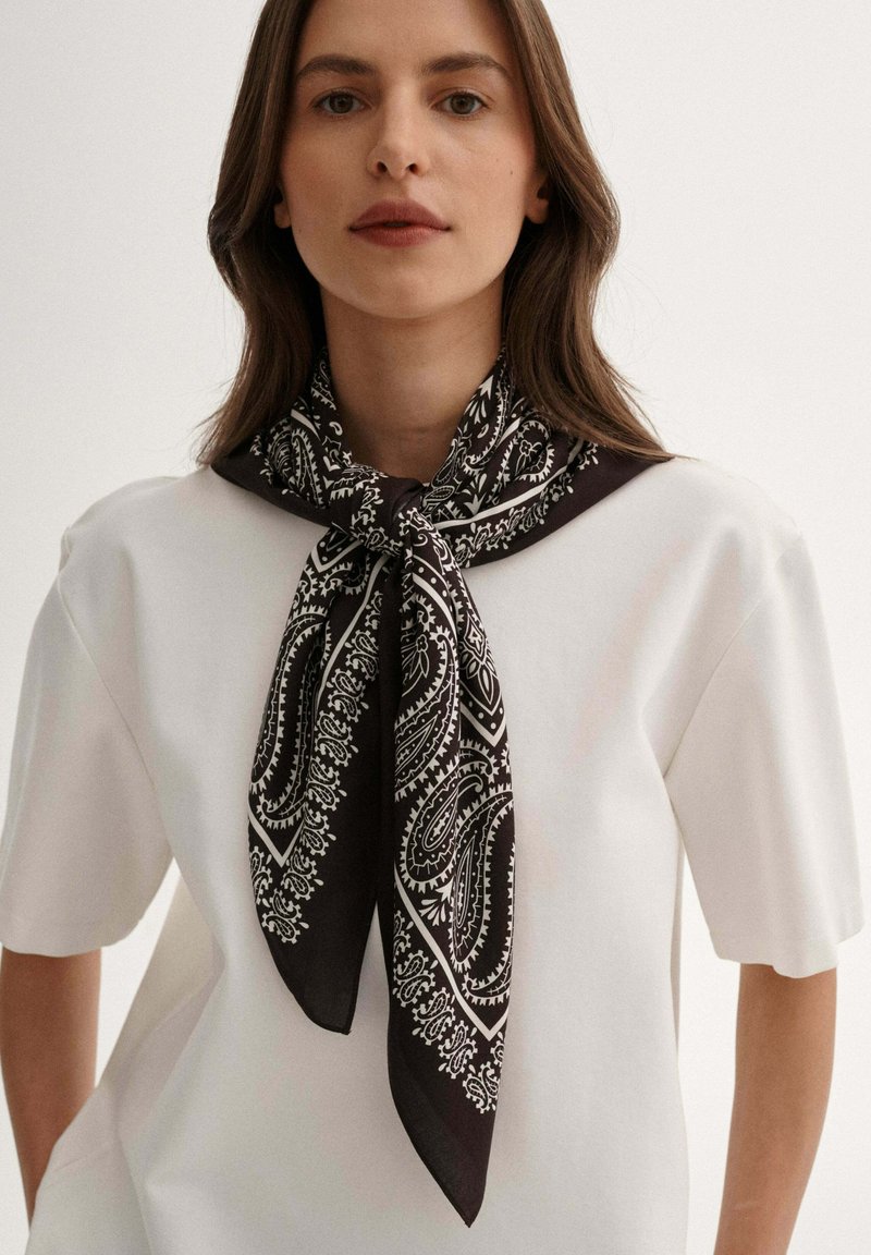 Czarny i biały jedwabny szalik w wzór paisley związany na szyi, kontrastujący z gładką, białą koszulą z krótkim rękawem. Miękka tekstura.