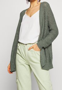 Žena v světle zelených cargo kalhotách, bílém topu s ramínky a otevřeném pleteném šedozeleném kardiganu, s jednou rukou v kapse na jednobarevném pozadí.