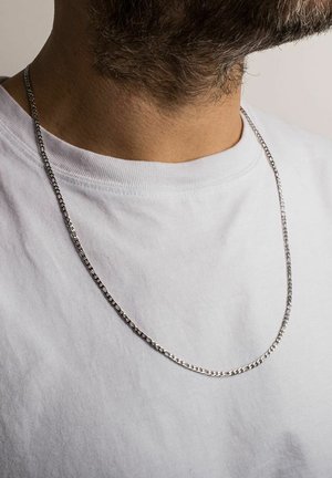 Collana in argento con un design piatto ed allungato. Presenta una texture liscia e riposa su una maglietta bianca semplice.