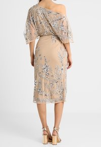 Beżowa sukienka z przezroczystą, haftowaną narzutą z srebrnymi kwiatowymi wzorami. Dopasowana spódnica, projekt jednego ramienia, zakończona grubymi sandałami na obcasie.