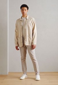 Giacca button-up beige chiaro con tasche frontali, abbinata a pantaloni a righe. Le sneakers bianche completano l'outfit. Texture lisce in tutto.