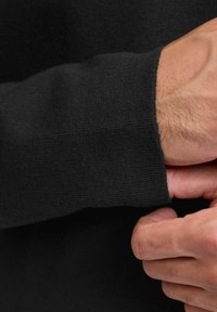 Egy kéz állítja a fekete, hosszú ujjú kötött pulcsi mandzsettáját, bemutatva a textúrát és a finom szövetnyúlást.