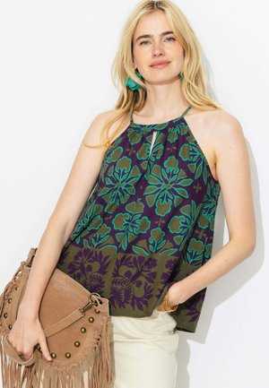 Femme blonde portant un haut dos nu floral vert et violet, un pantalon beige, des boucles d'oreilles turquoise, un bracelet en or, tenant un sac à main en daim beige à franges.