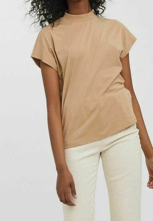 Femme portant un haut beige à manches courtes avec col montant et un pantalon blanc cassé, debout devant un arrière-plan clair uni.