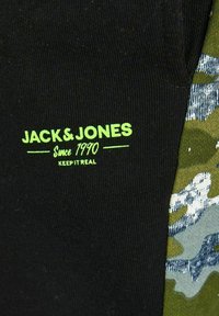 Sort stof med en ribbet tekstur, der har grøn tekst: "JACK & JONES Siden 1990 HOLD DET ÆGTE." Camouflage-mønster i nuancer af grøn og grå ved siden af.