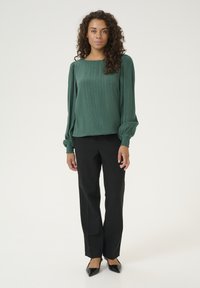 Grüne strukturierte Bluse mit langen Puffärmeln und Quadrat-Ausschnitt, kombiniert mit schwarzen maßgeschneiderten Hosen und schwarzen spitzen Schuhen.