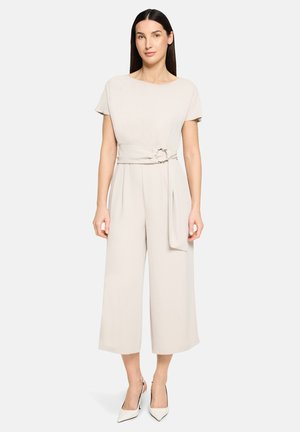 Femme aux cheveux raides et foncés portant une combinaison beige clair à manches courtes, jambes larges, et ceinture avec boucle métallique.