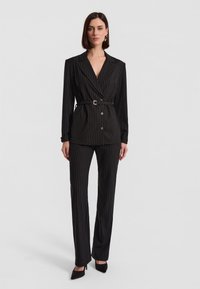 Czarny garnitur w prążki z podwójnym żakietem z paskiem w talii oraz dopasowanymi spodniami. Zawiera czarne szpilki.