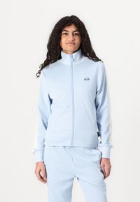 Lys blå zip-up hettegenser laget av myk stoff, med stående krage, subtilt logo og hvit-grønt stripete detaljer på ermene.