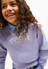Fioletowy sweatshirt z białym haftowanym napisem "TOMMY HILFIGER". Posiada jasnoniebieski wąski kołnierz pod spodem. Miękka tekstura i dopasowany krój.