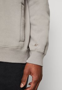 Hellgrauer Pullover mit gerippten Bündchen und Tasche. Ausgestattet mit strukturiertem Stoff und dezent gesticktem Logo in Dunkelbraun am Ärmelbündchen.