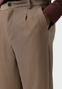 Pantalon beige côtelé avec un devant plissé. Il comporte une fermeture à bouton et des poches latérales. La texture du tissu est douce et structurée.