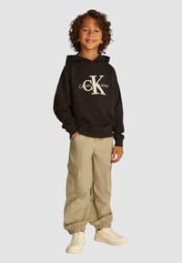 Felpa nera con cappuccio e logo bianco "Calvin Klein Jeans", abbinata a pantaloni cargo beige e sneaker bianche, con polsini elasticizzati.