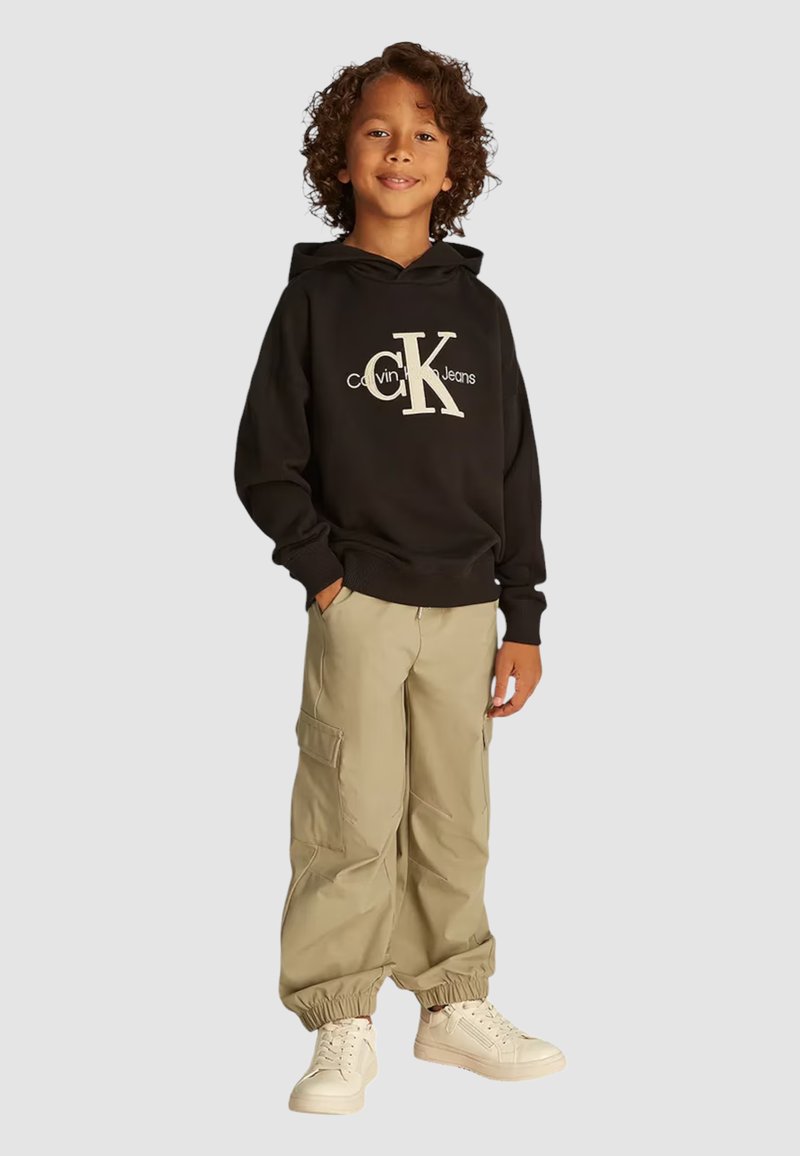 Felpa nera con cappuccio e logo bianco "Calvin Klein Jeans", abbinata a pantaloni cargo beige e sneaker bianche, con polsini elasticizzati.