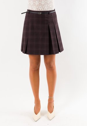 Donna che indossa una gonna plissettata a quadri scuri con cintura sottile, tacchi bianchi appuntiti e top in pizzo, in piedi su uno sfondo semplice.