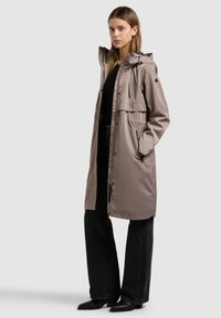 Naine, kes kannab beežikat kapuutsiga pikka vihmajope, musta topi, laia jalaga musti teksaseid ja musta teravate ninaosadega saapaid, seisab külje peal käed taskutes.