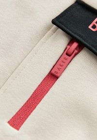 Lähellä punainen vetoketjun vetotuli, johon on kirjoitettu "BAKER", beige kankaalla ja mustalla märkinnän reunalla, jossa on osittainen punainen tekstitys.