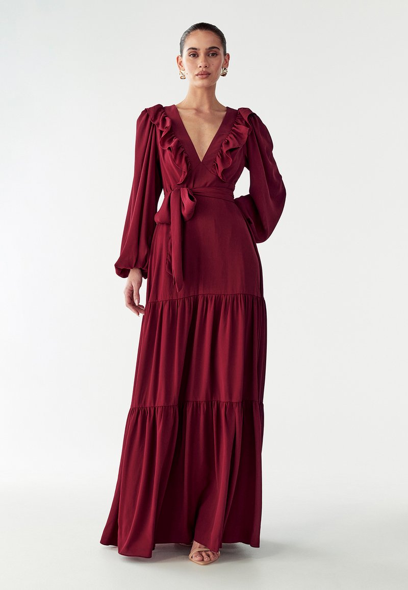 Burgundi spalvos maxi suknelė su giliu V formos iškirpimu ir raukiniais. Ilgos rankovės, surišta talija ir sluoksniuota sijoną sukuria plazdančią siluetą.
