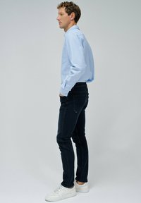 Camisa de botones azul claro, jeans oscuros de corte slim y zapatillas blancas. El modelo está de lado, mostrando un bolsillo en la camisa y líneas limpias.