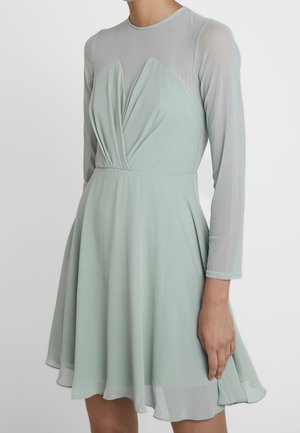 Femme portant une robe vert pâle à manches longues transparentes et un corsage drapé en forme de V, debout devant un fond clair uni.