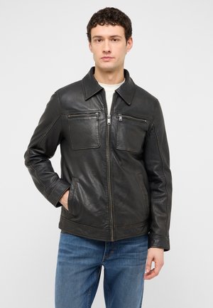Man wearing a black leather jacket with zipped chest pockets and blue jeans, standing against a plain light background.