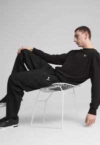 Se lleva un sudadera negra y pantalones cargo, con un pequeño logo; el modelo está sentado en una silla de alambre blanca, se ven unas zapatillas negras.