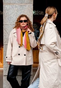 Beige double-breasted coat, black leather pants, layered colored scarves (pink and yellow) and small black sunglasses. Gray scarf on hair.