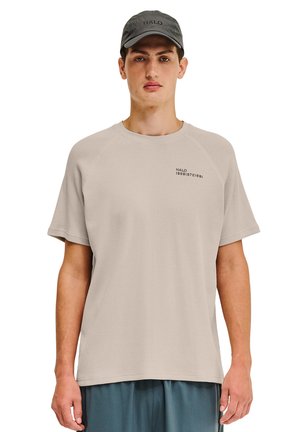 Jasno-beżowy, teksturowany t-shirt z krótkim rękawem, z okrągłym dekoltem i małym czarnym logo po lewej stronie. Nosi czarną czapkę.