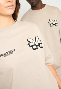 Camisetas de algodón beige con cuello redondo, que presentan un logotipo impreso en negro "MULTIPLY APPAREL" y un diseño bordado de "M" en el pecho.