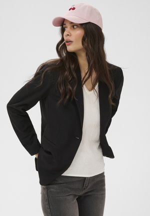 Mujer con cabello largo y castaño lleva una gorra rosa con bordado de cerezas, blazer negro, blusa blanca y vaqueros grises, mirando hacia un lado.