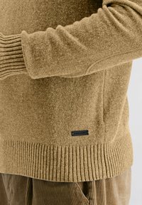 Close-up van een persoon in een tan gebreide trui met geribde manchetten en zoom, en tan corduroy broek, waarbij een klein merkembleem op de trui zichtbaar is.