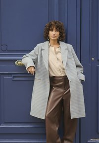Cappotto doppiopetto in blu chiaro e motivo houndstooth crema, indossato sopra una camicia beige con bottoni e pantaloni marroni in ecopelle.