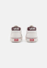 Baskets montantes Vans blanches avec un empiècement talon bordeaux, arborant le logo "VANS" au dos et une étiquette classique en caoutchouc rouge et blanc.