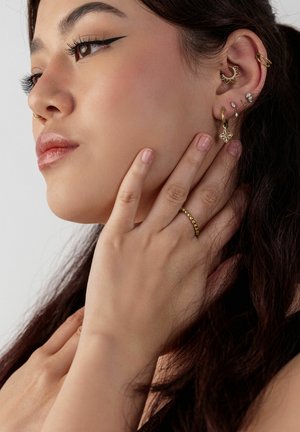 Junge Frau mit langen dunklen Haaren berührt ihren Hals, trägt mehrere goldene Ohrringe, einen Nasenring und einen goldenen Perlenring am Finger.