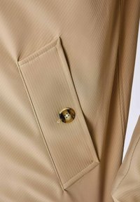 Tela beige con una fina textura acanalada, que presenta un bolsillo y un botón redondo con un patrón en la solapa, mostrando detalles de diseño sutiles.