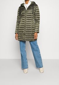 Persoon in olijfgroene gewatteerde hoodie-jack over een witte top en blauwe wijde jeans, staande tegen een plain witte achtergrond.