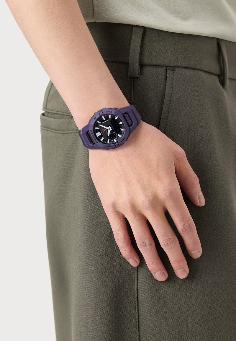 Montre en caoutchouc violette avec un cadran analogique noir, des index horaires blancs et des accents rouges, portée sur une main reposant sur une poche de pantalon verte.