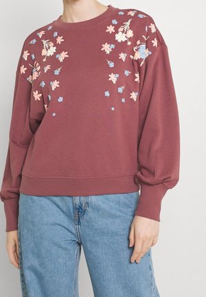 Persoon draagt een mauve sweatshirt met roze en blauwe bloemrijke borduursels op de schouders en de bovenkant van de borst, gecombineerd met lichtblauwe spijkerbroek.