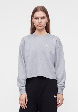 Giovane donna con capelli lisci castano chiaro indossa una felpa corta grigio chiaro e pantaloni neri con loghi bianchi sul petto sinistro e sulla coscia.