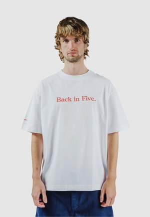 Jonge man met golvend lichtbruin haar, gekleed in een wit T-shirt met de tekst "Back in Five." en donkerblauwe broek, staand tegen een effen achtergrond.