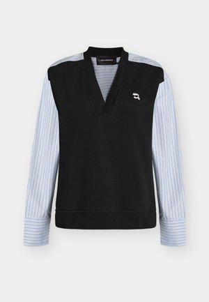Top noir sans manches à col en V superposé sur un haut à manches longues bleu et blanc rayé, avec un petit logo blanc sur la poitrine gauche.