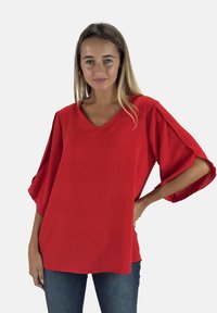 Blusa rossa a maniche corte con scollo a V e tessuto strutturato. Vestibilità larga con maniche ampie, abbinata a jeans di denim blu.