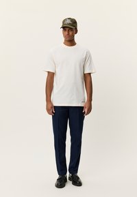 Λευκό βαμβακερό T-shirt με κοντά μανίκια, συνδυασμένο με μπλε σκούρο παντελόνι και μαύρα παπούτσια. Καπέλο ελιάς με κεντημένη λεπτομέρεια στο μπροστινό μέρος.