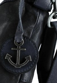 Borsa in pelle nera con un'etichetta circolare con un emblema di ancora in metallo argentato. Sono visibili dettagli cuciti e una texture liscia.