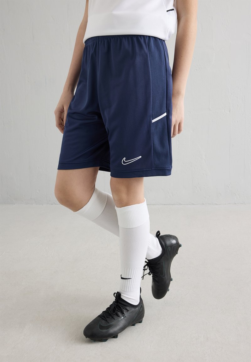 Persona tērpusies tumši zilos sporta šortos, baltās ceļgalu zeķēs ar melnu svītru un melnos futbola apavos, stāv ar vienu kāju pāri otrai.